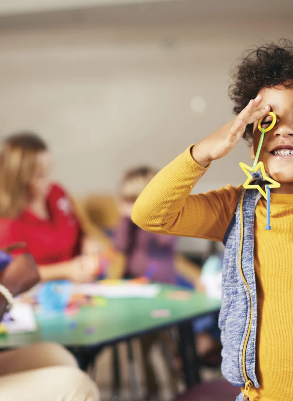 A young African-American child is smiling and playing with toys or craft supplies while Y staff smile and play with other children in the background. Find your Y