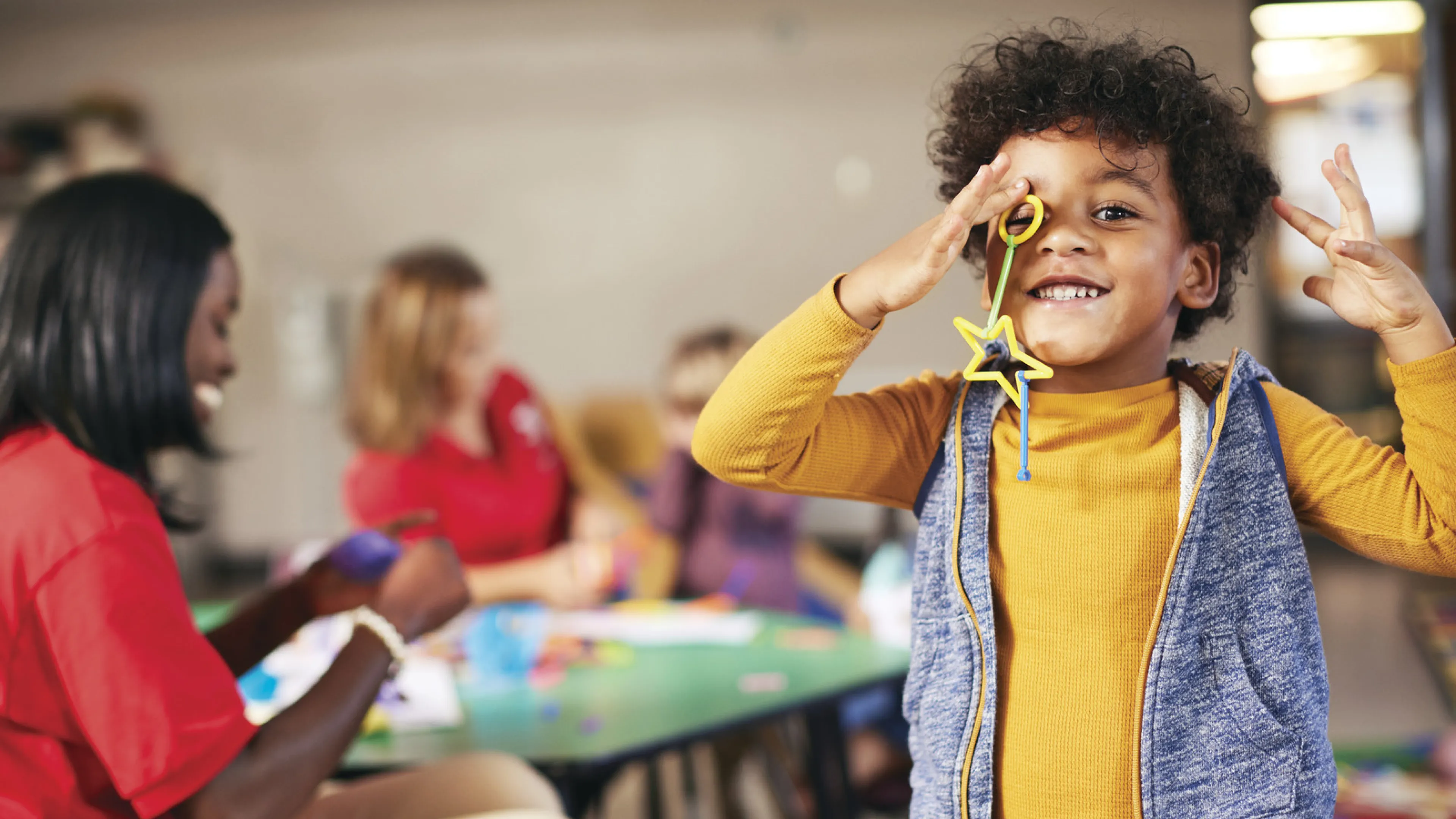 A young African-American child is smiling and playing with toys or craft supplies while Y staff smile and play with other children in the background. Find your Y