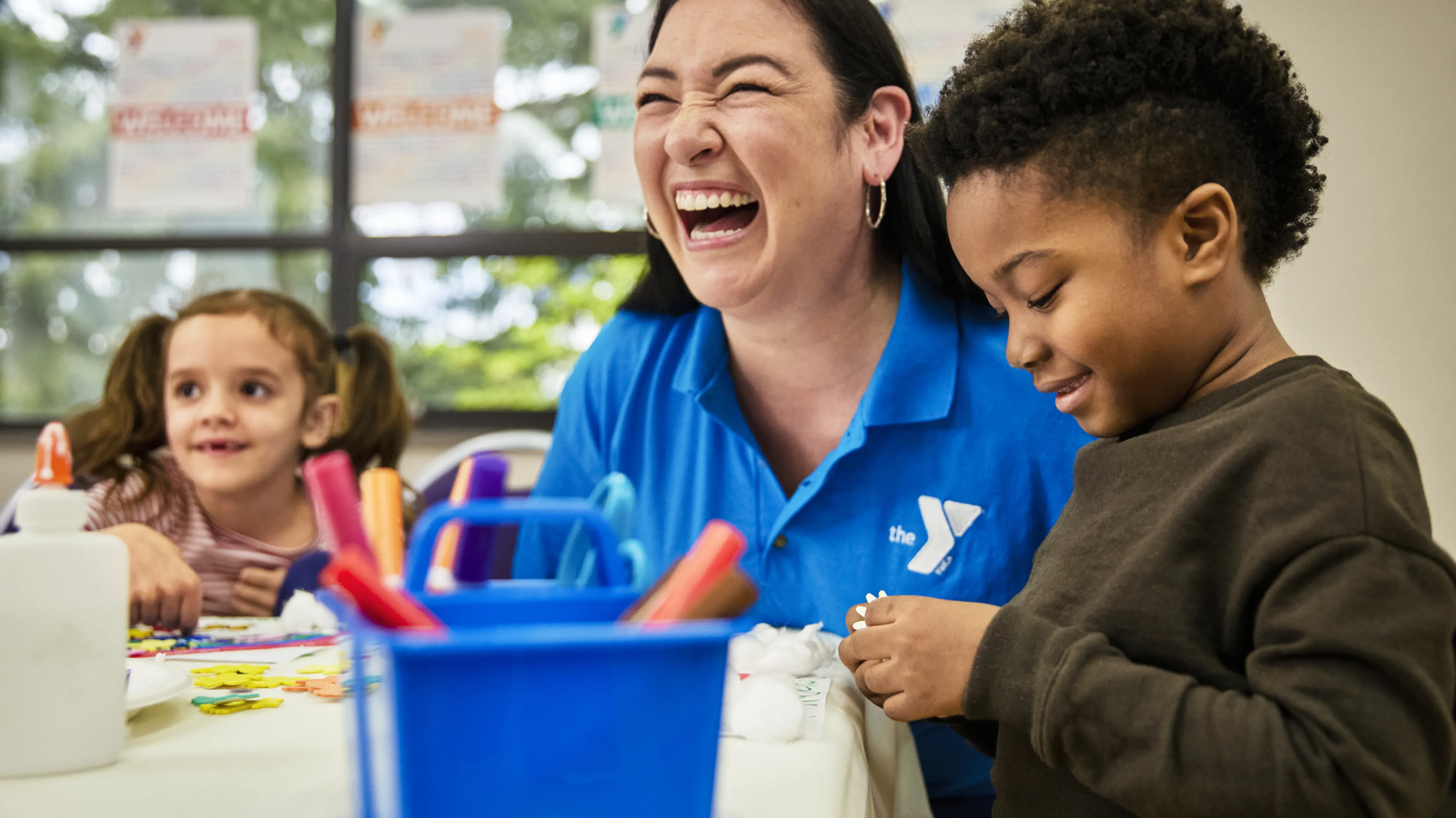 After school a Y staff member laughs and smiles while helping two young children with an art project that involves rainbows and markers; 2022 Afterschool Staff Recruitment