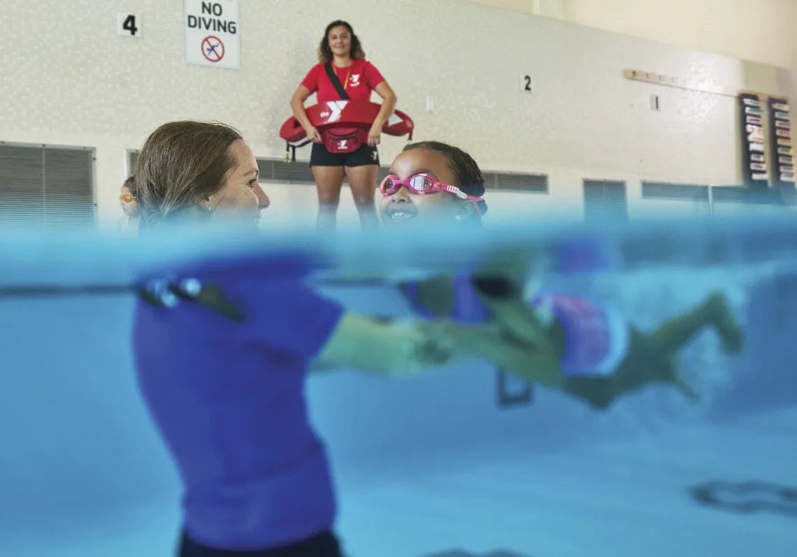 A YMCA swim instructor helps a school age child learn to swim during her swim lesson in the indoor pool as the lifeguard watches.