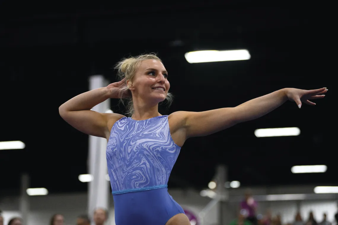 A teenage female gymnast in a blue outfit is shown striking a pose with her arms to the right on the floor mat during her routine while spectators watch at the YMCA National Gymnastics Championships.